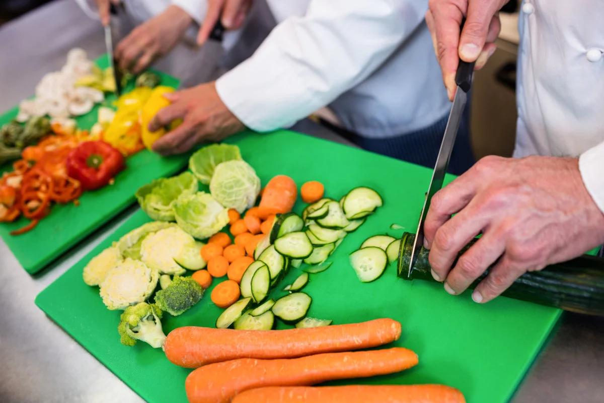 Diverse mani di cuochi tagliano e preparano una varietà di verdure fresche, tra cui carote, cetrioli e peperoni, disposte su taglieri verdi.
