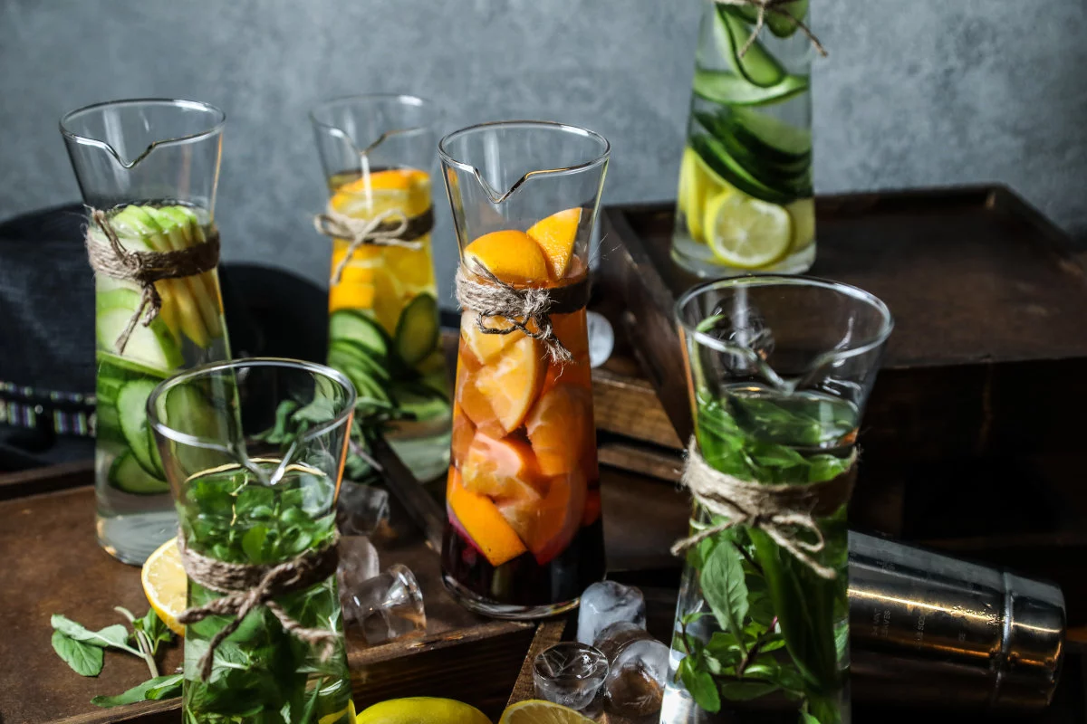Diverse caraffe di vetro contenenti acqua aromatizzata con fette di frutta come cetrioli, arance e limoni, oltre a foglie di menta, sono ornate con spago e poste su un ripiano di legno con cubetti di ghiaccio sparsi e uno shaker in metallo.