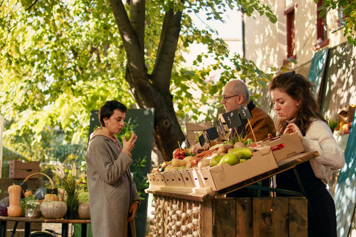 In un mercato allaperto sotto gli alberi, tre persone interagiscono attorno a un banco ricco di frutta e verdura fresca, inclusa una donna che osserva attentamente una mela verde.