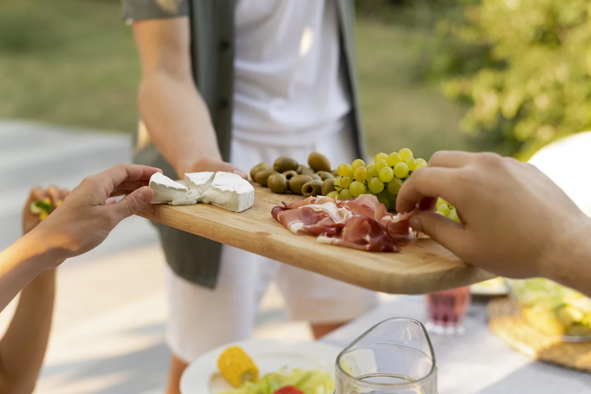 Mani afferrano formaggio, olive, uva e prosciutto da un tagliere di legno servito allaperto.