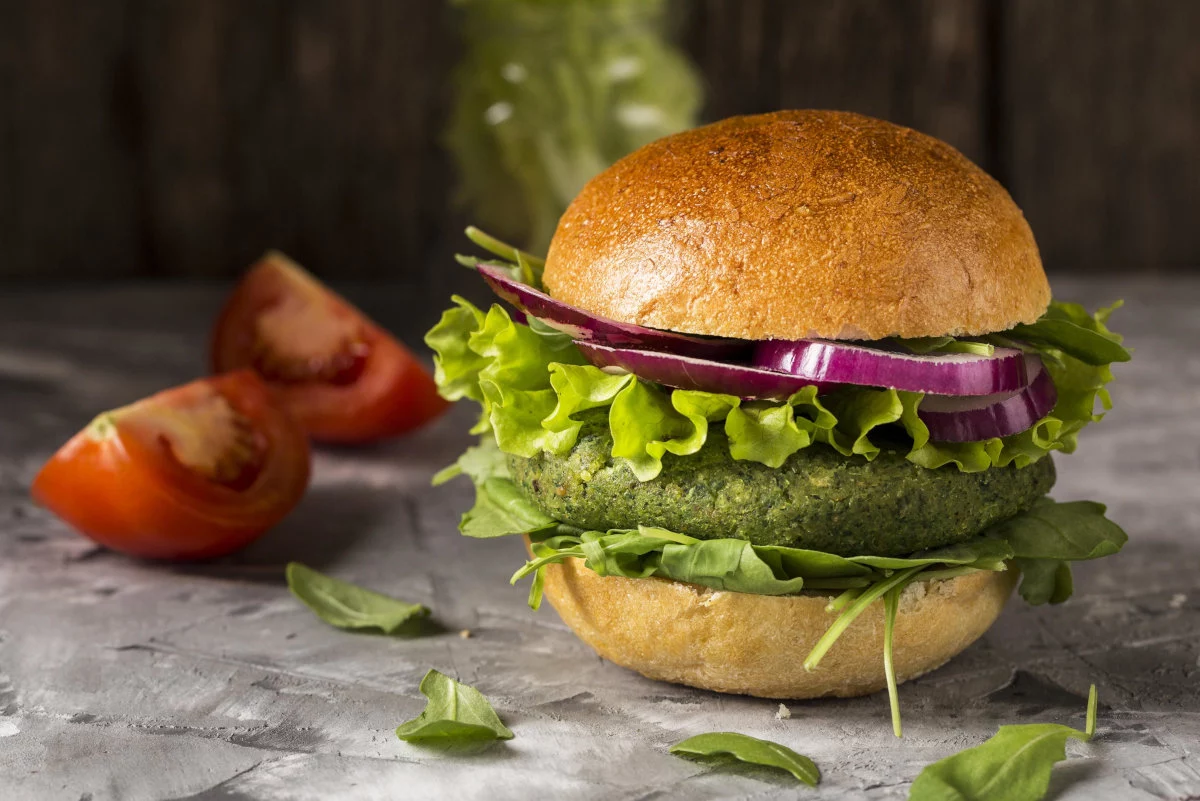 Un hamburger vegetariano con polpetta verde, lattuga e cipolla rossa è appoggiato su una superficie grigia accanto a due spicchi di pomodoro.