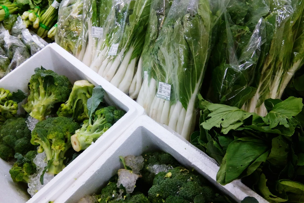 Limmagine presenta broccoli verdi freschi in contenitori di polistirolo con ghiaccio e diverse varietà di verdure a foglia verde, alcune avvolte singolarmente in plastica.