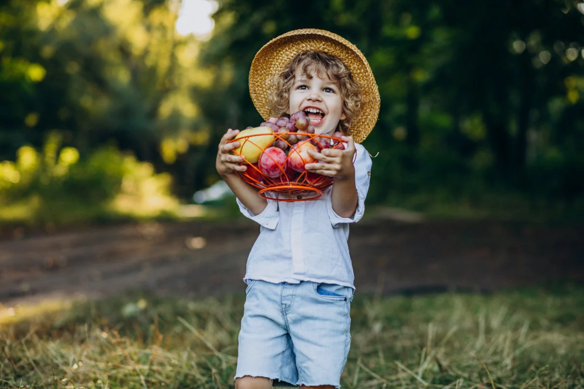 Un bambino riccio con un cappello di paglia sorride felicemente con la bocca aperta, reggendo un cesto ricolmo di frutta assortita in un ambiente naturale.