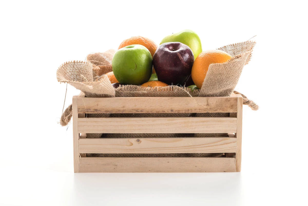 Una cassa di legno ripiena di mele verdi e rosse e arance, adagiate su tela di iuta, è visibile su sfondo bianco.