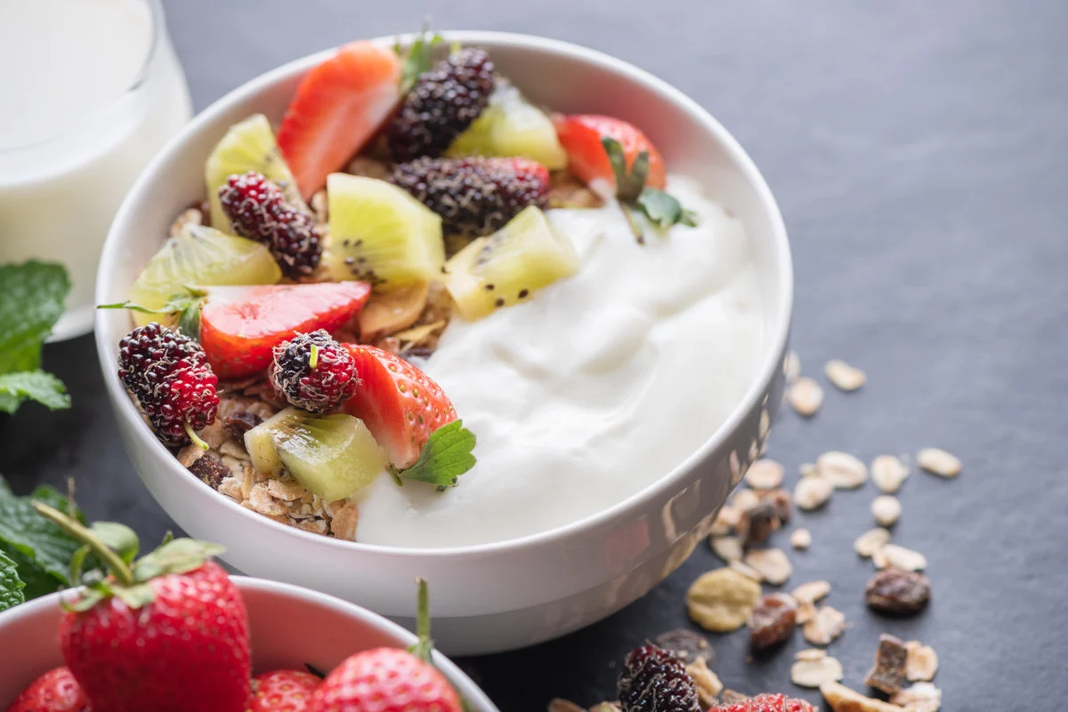 Limmagine raffigura una ciotola di yogurt guarnita con fragole, kiwi, more di gelso e cereali sparsi, con altre fragole e una brocca di latte visibili sullo sfondo su una superficie scura.