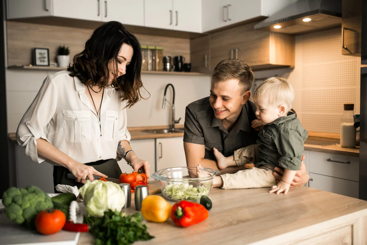 Una famiglia è intenta a preparare un pasto in una cucina moderna, con la donna che taglia verdure fresche mentre luomo sorride tenendo un bambino piccolo seduto sul bancone.