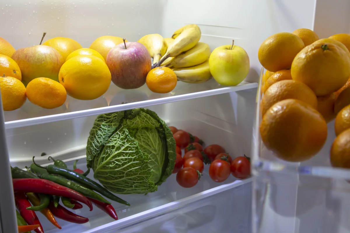 Un frigorifero aperto rivela ripiani pieni di frutta e verdura fresca, tra cui mele, banane, arance, limoni, cavolo, pomodori e peperoncini.