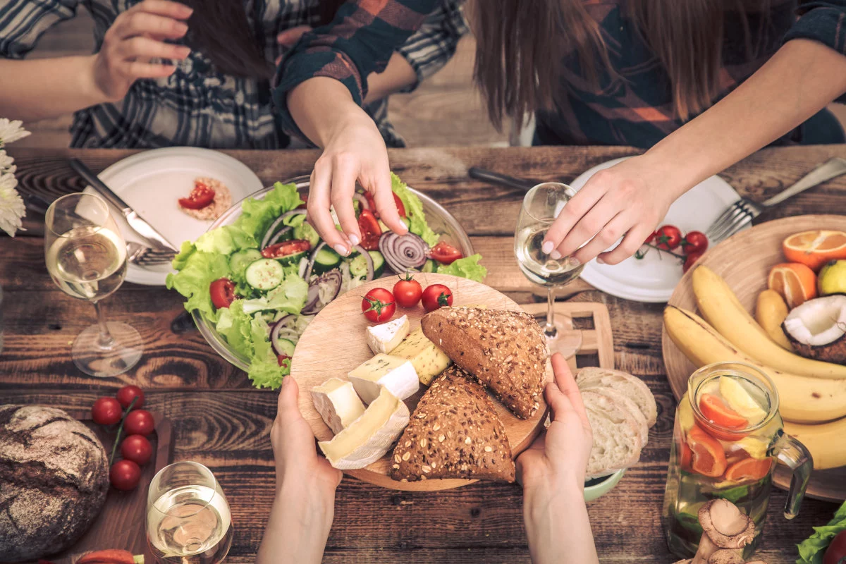 Durante un pasto conviviale, diverse mani si servono da una tavola di legno imbandita con insalata, pane, formaggi, frutta, pomodori e bicchieri di vino.
