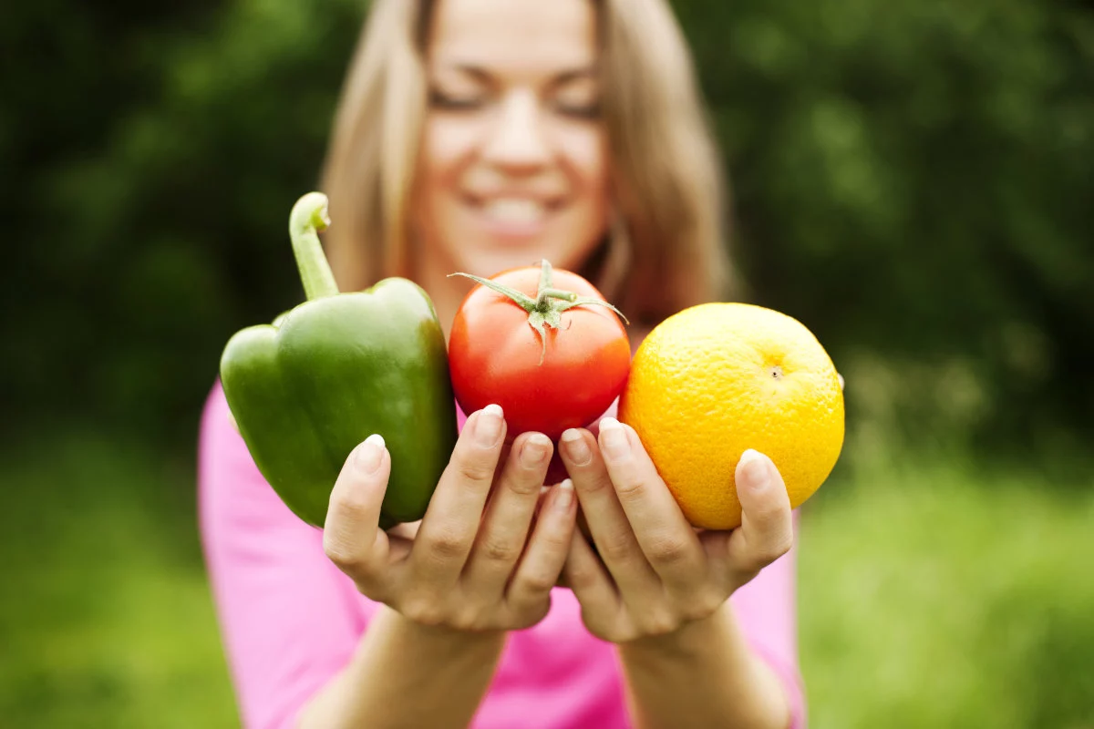 Le mani di una donna, con il volto sorridente sfocato, mostrano un peperone verde, un pomodoro rosso e un agrume giallo su uno sfondo naturale.