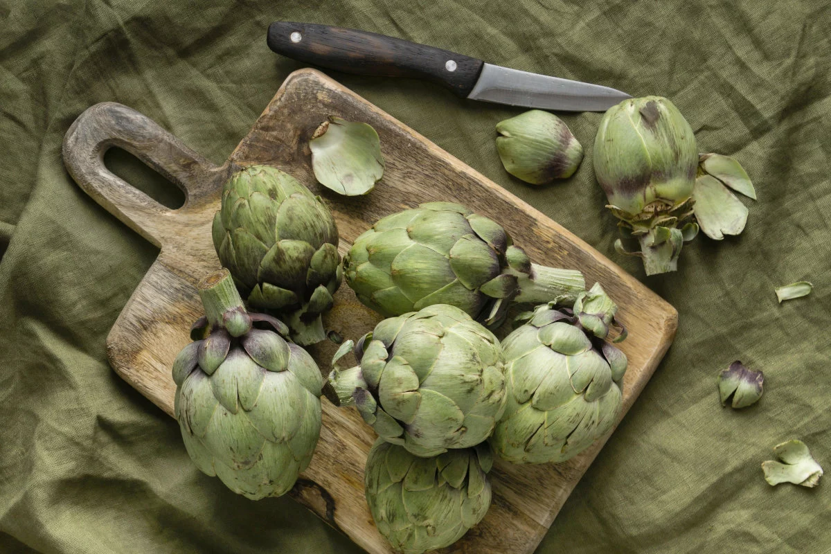 Diversi carciofi verdi freschi sono disposti su un tagliere di legno e su un panno verde, con un coltello e alcune foglie sparse.