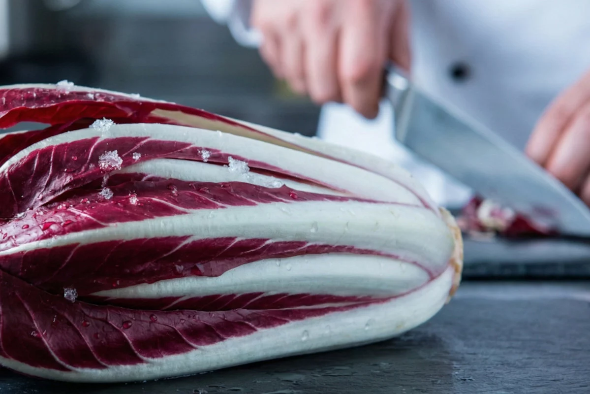 Un radicchio bicolore con goccioline dacqua e piccoli cristalli sulle foglie è in primo piano, mentre sullo sfondo un cuoco taglia delle verdure con un coltello.
